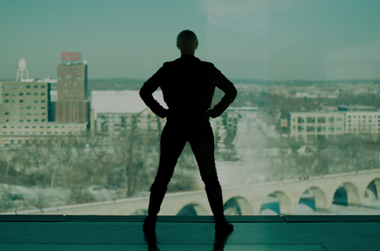 Justine stands in a super hero pose, hands on hips, silhouetted against the Minneapolis skyline.